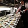 Plates with open-faced sandwiches are being garnished by a chef in a professional kitchen. The sandwiches are arranged neatly on a stainless steel counter under bright lights.