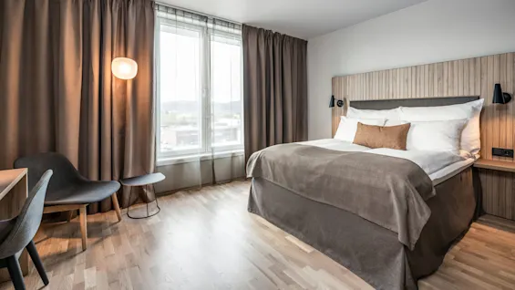 En seng med brunt sengetøj står under en paneleret hovedgavl. En moderne stol og lampe er i nærheden i et blødt belyst rum med store vinduer og brune gardiner.