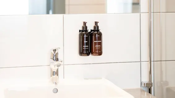 Två bruna pumpflaskor märkta "BODY LOTION" och "SOAP" är monterade på en kaklad badrumsvägg ovanför ett vitt handfat med en kromkran; en delvis öppen dörr syns i bakgrunden.