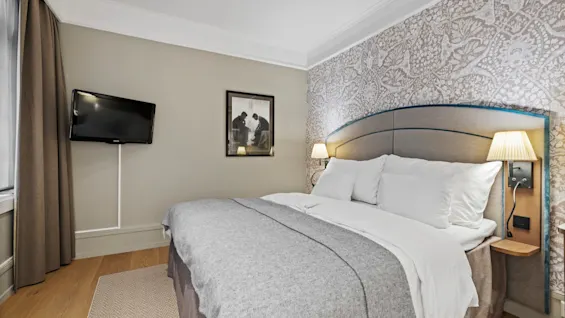 A comfortable hotel room featuring a large bed with white linens, patterned wallpaper, a wall-mounted television, and a framed artwork.
