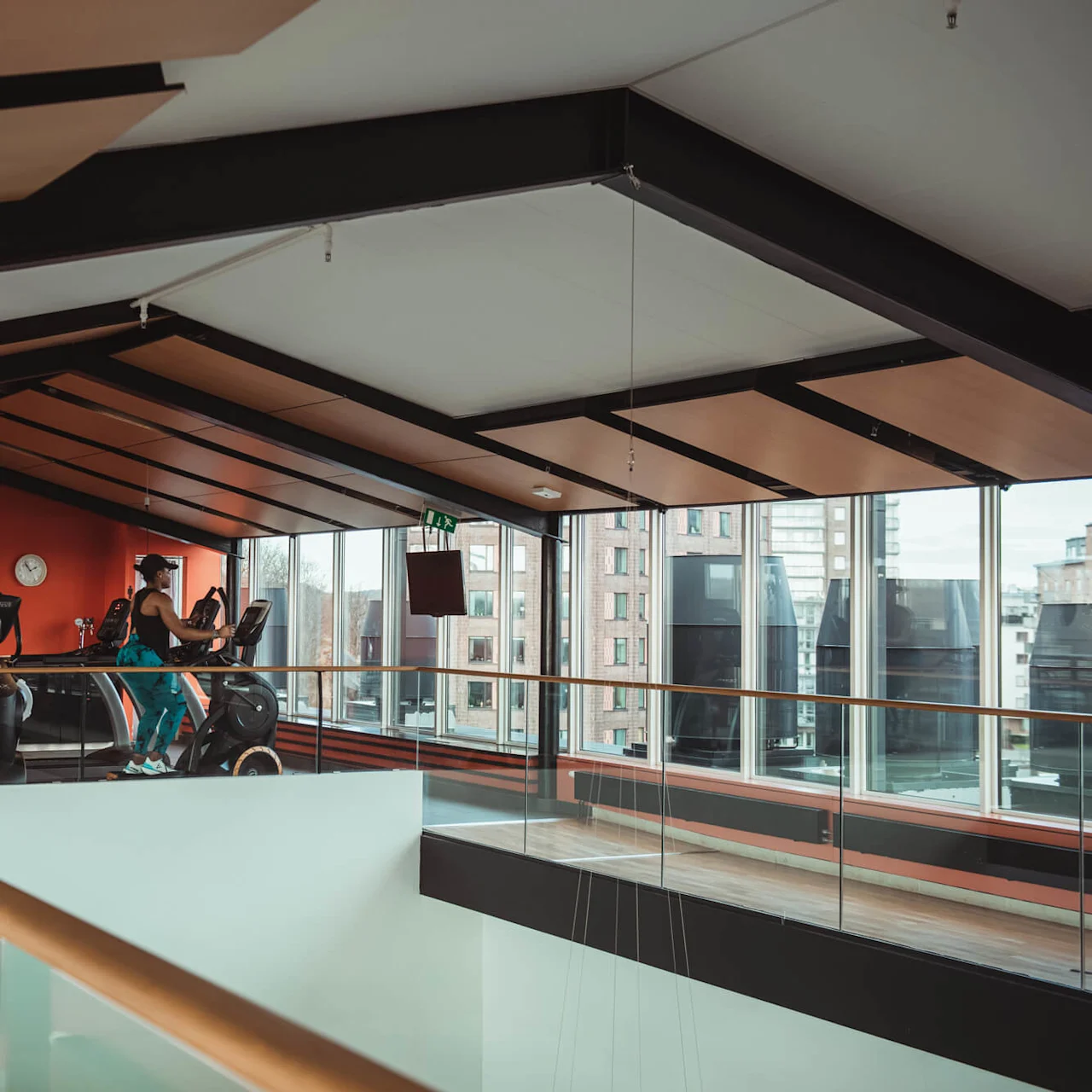 A person exercises on a treadmill inside a modern gym with large windows and wooden ceilings. A clock and various exercise machines are visible against red walls.