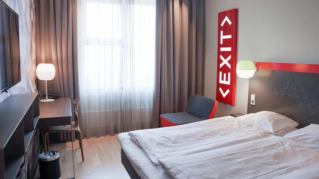 A minimalistic hotel room with a bed, chair, TV, and a lamp. An illuminated "EXIT" sign adds a quirky touch against a gray wall.