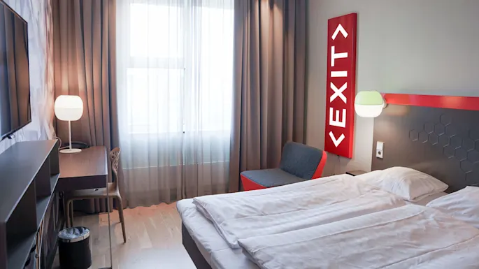 Et minimalistisk hotellrom med en seng, stol, TV og en lampe. Et opplyst "EXIT"-skilt tilfører en quirky detalj mot en grå vegg.