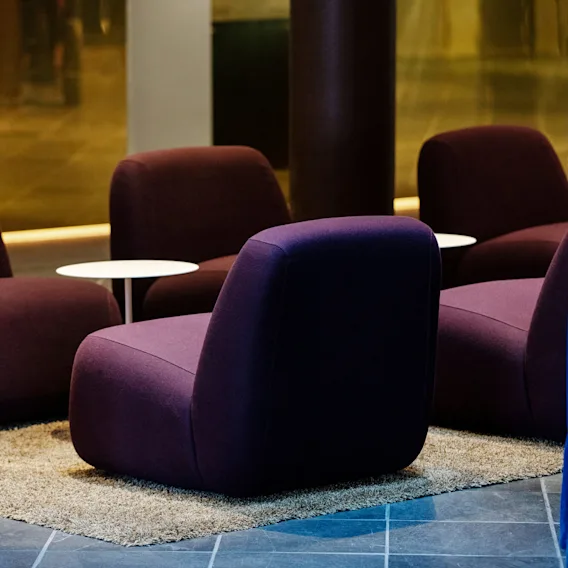 Purple lounge chairs sit on a beige carpet, paired with small white tables. They are surrounded by dark flooring and blue curtains, creating a cozy seating area.