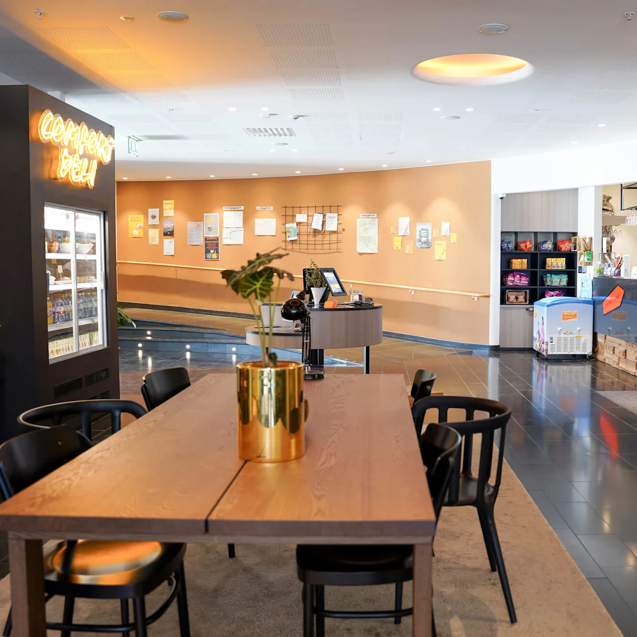 Caféinteriør med trebord, sorte stoler og en disk. Neonskilt med "Corner's Deli", planter som dekor og kunder tilstede.