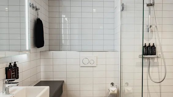 Puhdas, moderni kylpyhuone, jossa on valkoiset laatat, pesuallas, wc kaksoishuuhtelupainikkeilla ja suihku tuotteineen.