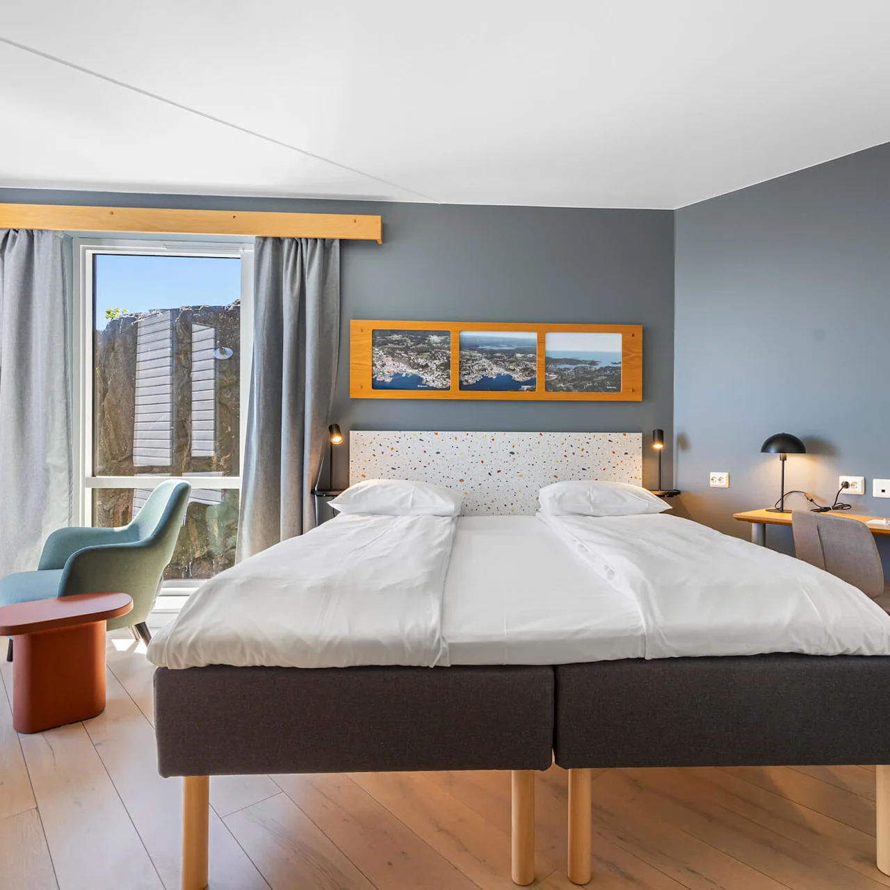 A modern bedroom with twin beds, a desk, chair, and artwork, illuminated by natural light from a window.