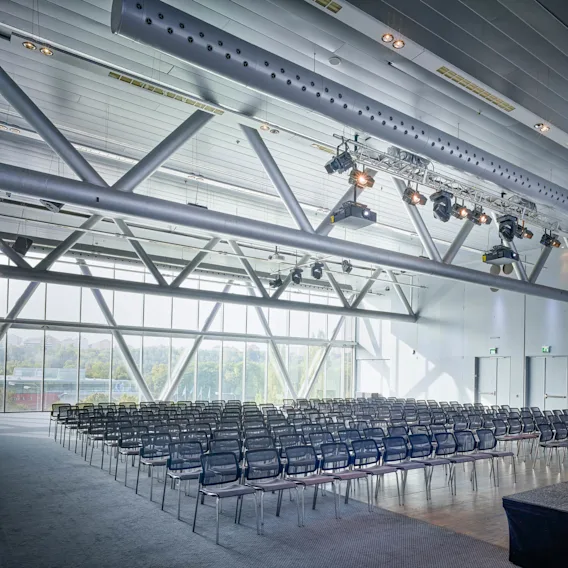 Rows of empty black chairs face a stage in a modern, light-filled conference room with large windows and overhead lighting. Visible greenery outside the windows enhances the setting.