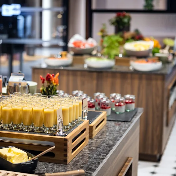 Glass med oransje smoothie står på et trebrett, omgitt av en buffé med ulike matvarer. Tekst inkluderer "gul smoothie" og "rød smoothie."