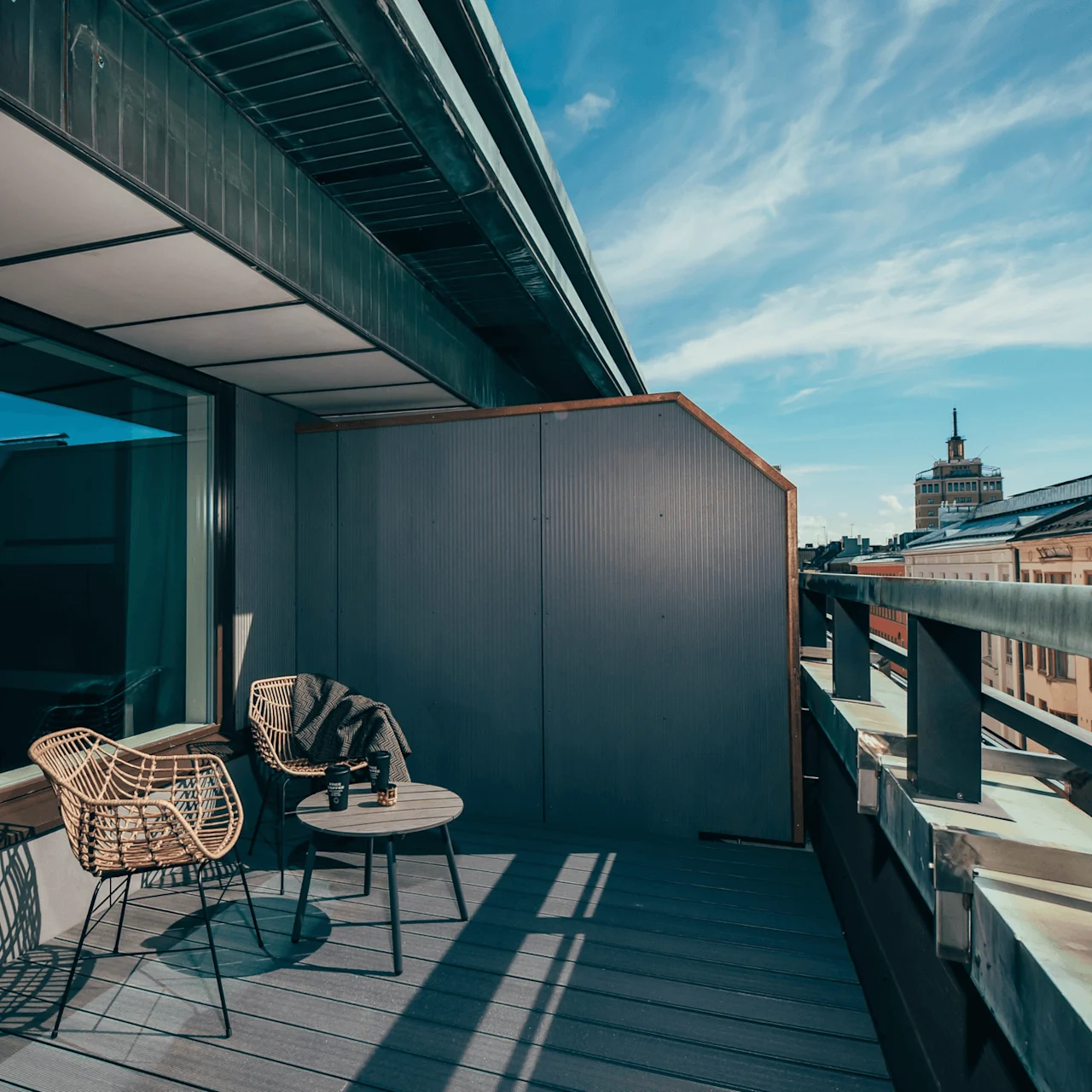 To rottingstoler og et lite rundt bord står på en moderne takterrasse under en klar blå himmel. Smale bygninger og et fjernt tårn er synlige i horisonten.