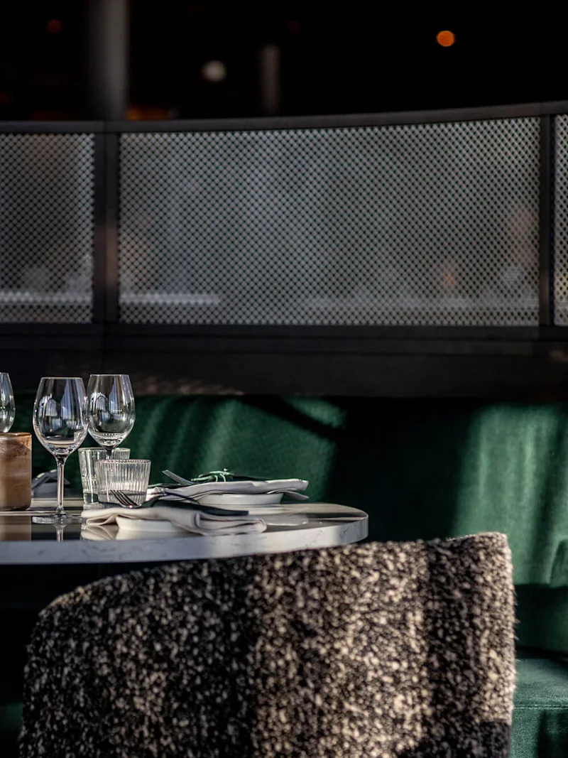 Pyöreä ruokapöytä, jossa on viinilasit, lautasliinat ja aterimet; ympäröity pehmeillä tuoleilla himmeästi valaistussa, viihtyisässä ravintolassa.