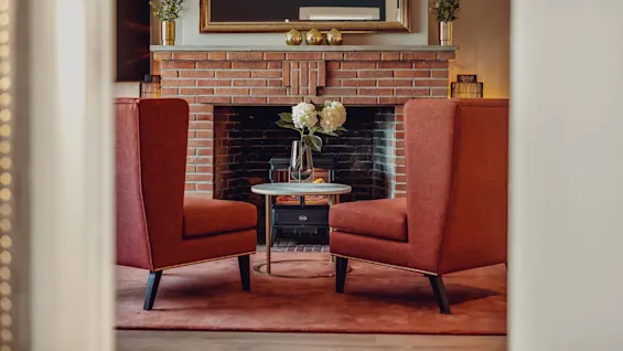 Two terracotta armchairs flanking a round table by a brick fireplace with decorations, in a cozy living room setting.