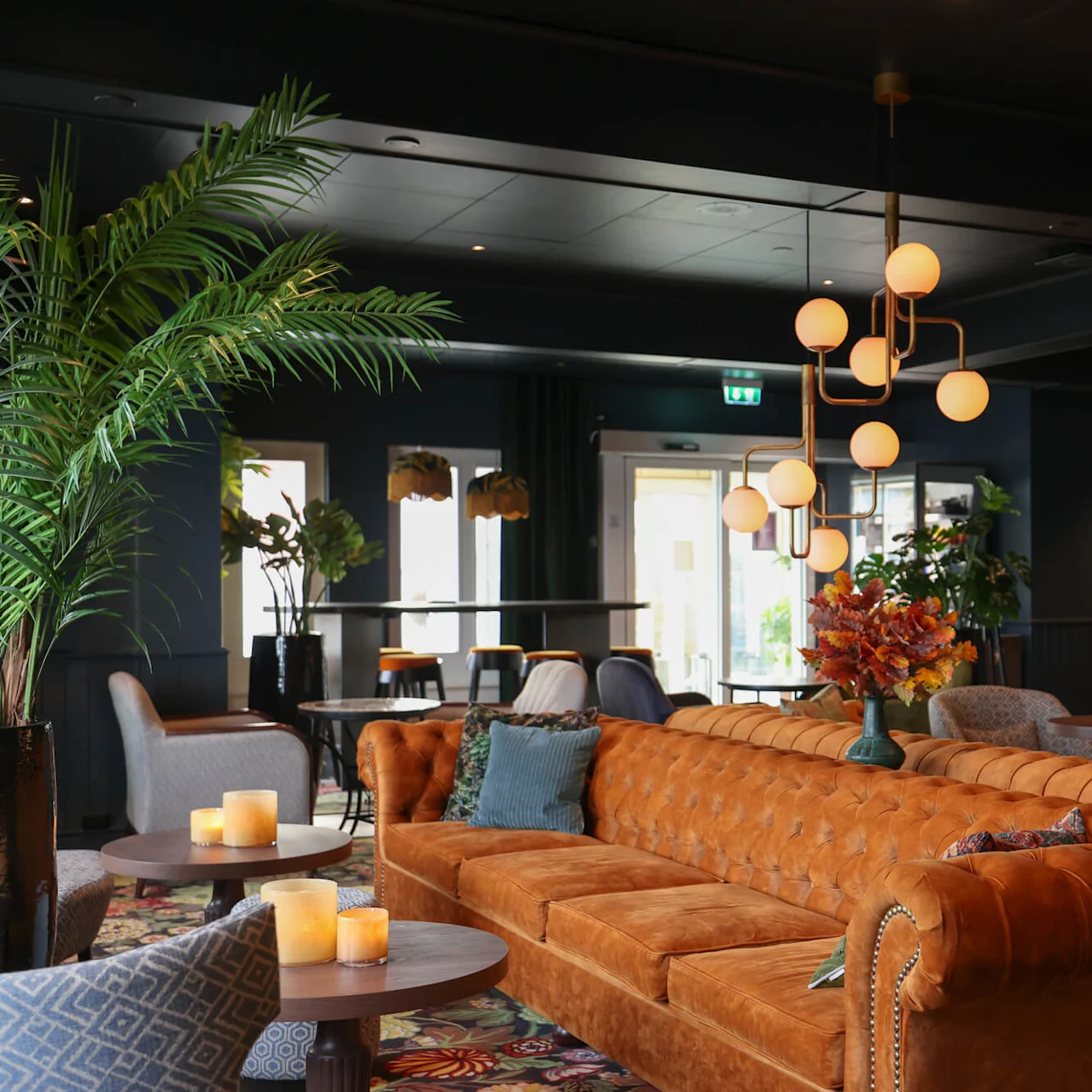 A brown leather couch adorned with cushions sits under a modern chandelier, surrounded by potted plants, chairs, and tables with candles in an elegantly decorated lounge.