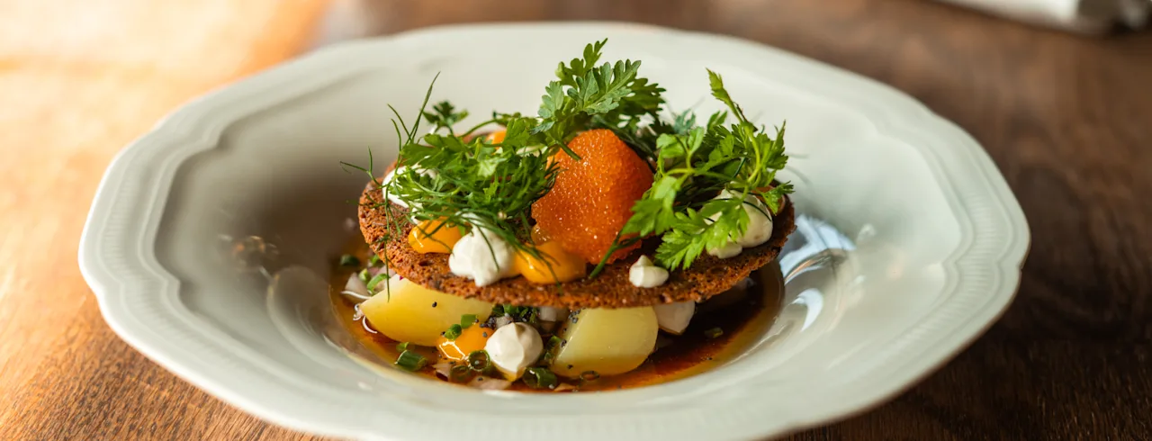Gourmet-annos tuoreilla yrteillä, oranssinvärisellä ruoalla ja perunoilla, tyylikkäästi tarjoiltuna valkoisella lautasella puupöydällä.