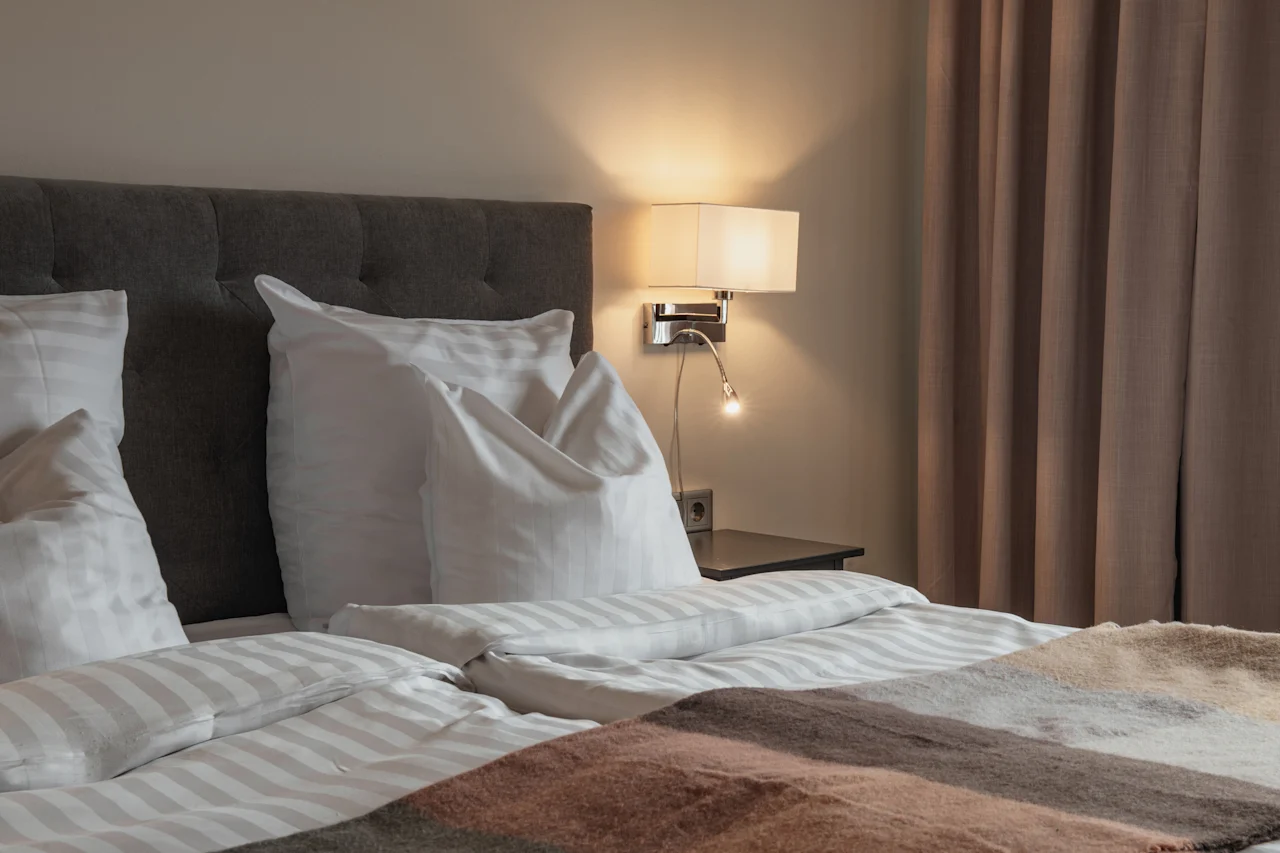 En komfortabel hotelseng med sprøde hvide lagner og et varmt tæppe, oplyst af en blød væglampe, klar til hvile.