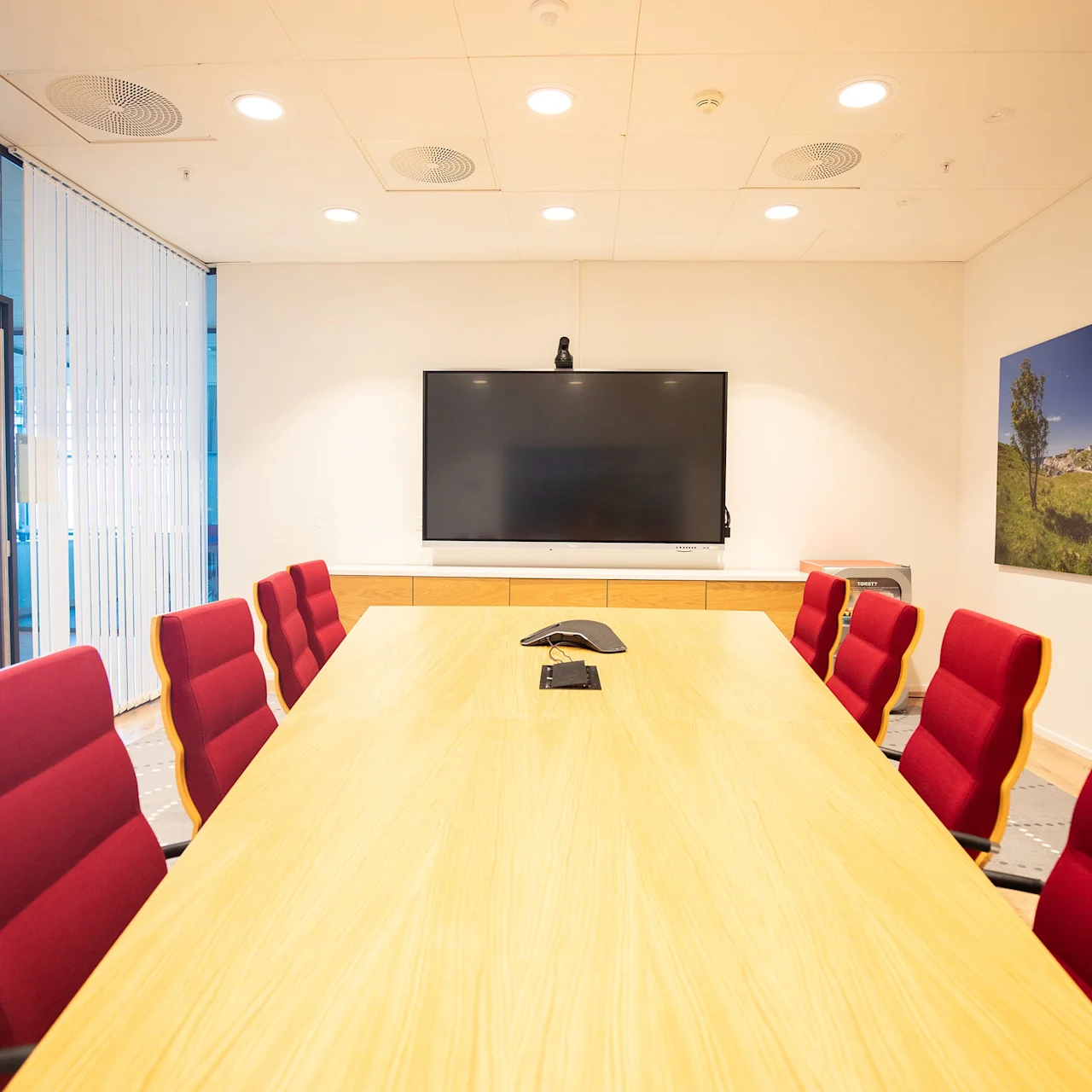 Et rektangulært konferansebord med røde stoler står i et møterom. En stor skjerm er montert på veggen, med et landskapsbilde ved siden av. Lys belysning over hodet.