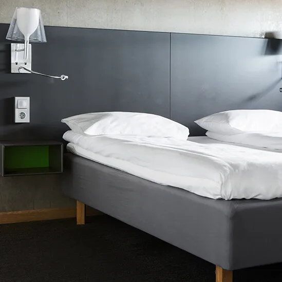 Sängyn päällä yksi siisti kahden hengen sänky valkoisin lakanoin, tummanpuhuva pääty. Kaksi seinävalaisinta ylhäällä, vieressä verhot modernissa, minimalistisessa makuuhuoneessa.