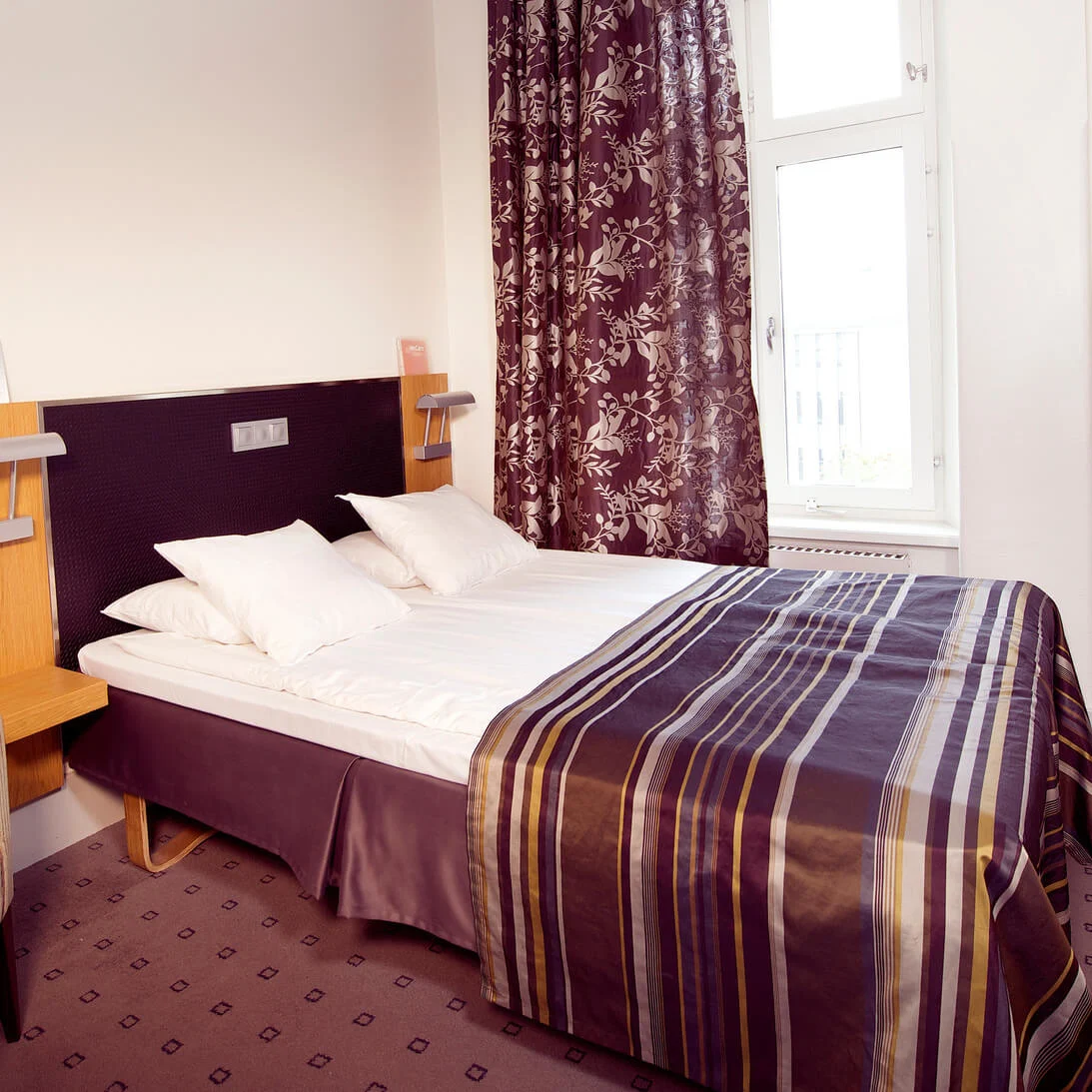 A neatly made bed in a hotel room with a striped blanket, flanked by a chair and a television on the wall.