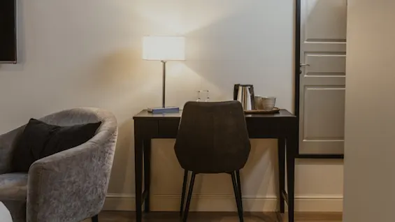 Et behageligt skrivebordsområde på et hotelværelse med en lampe, stol og kaffestation, ved siden af en plys lænestol.