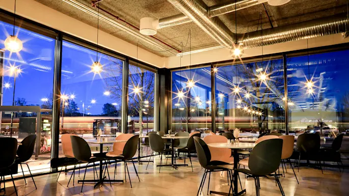 A modern café featuring empty tables and chairs, illuminated by hanging lights. Large glass windows reveal a street scene with passing vehicles and snow-covered trees outside.