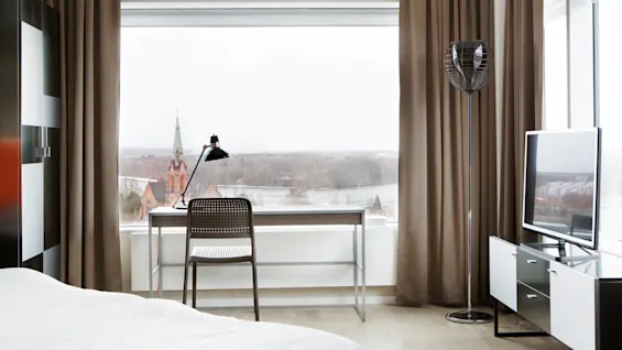 Et skrivebord med lampe og stol vender mod et stort vindue med udsigt over en fjern rød teglstenskirke. Rummet har en seng, gulvlampe og tv i moderne stil.