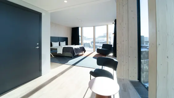 A modern bedroom features a neatly made bed with grey bedding, facing large windows letting in bright sunlight. The room includes a black door, chairs, and light-colored wooden walls.