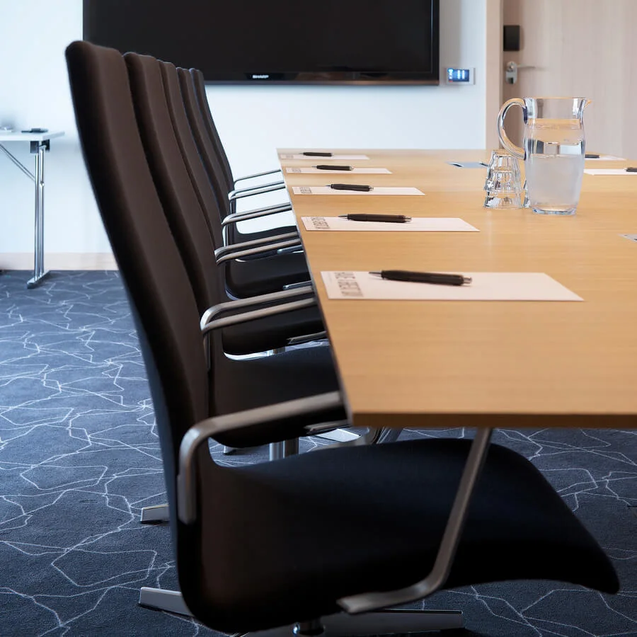 En rad svarta stolar står vid ett konferensbord med anteckningsböcker och pennor, framför en stor väggmonterad skärm i ett modernt mötesrum. En kanna med vatten och glas står på bordet.