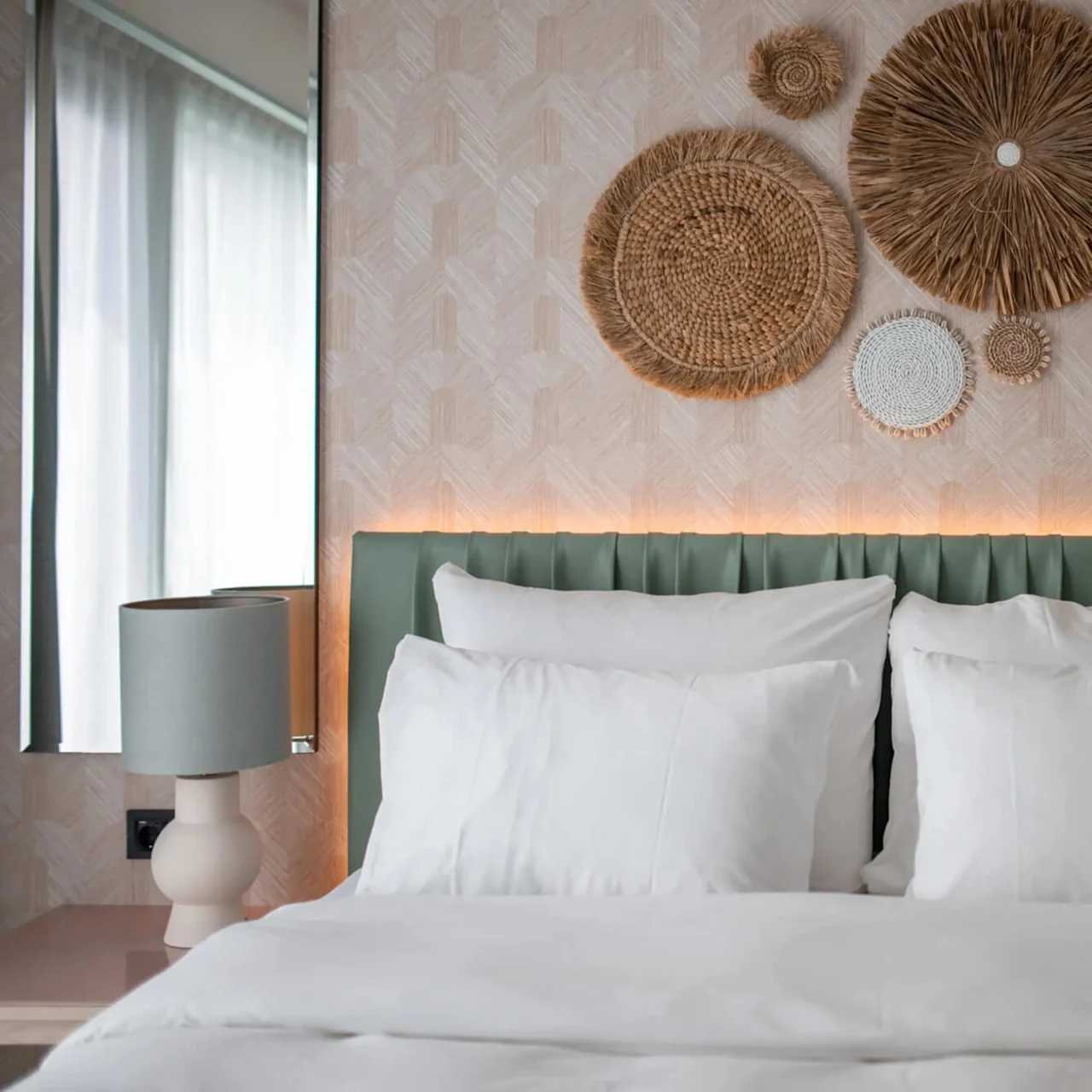 A neatly made bed with white pillows and a green headboard, flanked by lamps, under a woven circle wall art. Soft light glows behind the headboard. 