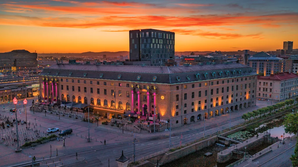 Ett stort hotell i klassisk stil upplyst i rosa och gult under en färgstark solnedgång i staden.