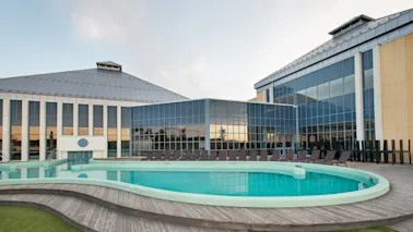 En cirkulär utomhuspool omges av trädäck, med solstolar placerade runt om. Poolen ligger bredvid en stor modern byggnad med glaspaneler.