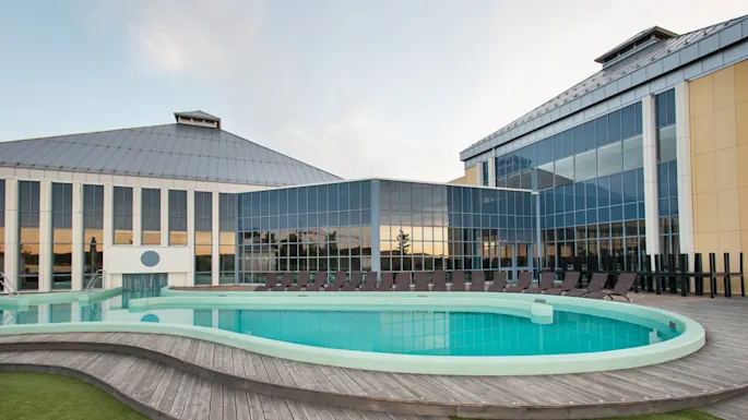 A circular outdoor swimming pool is surrounded by a wooden deck, with loungers positioned around it. The pool is adjacent to a large modern building with glass panels.