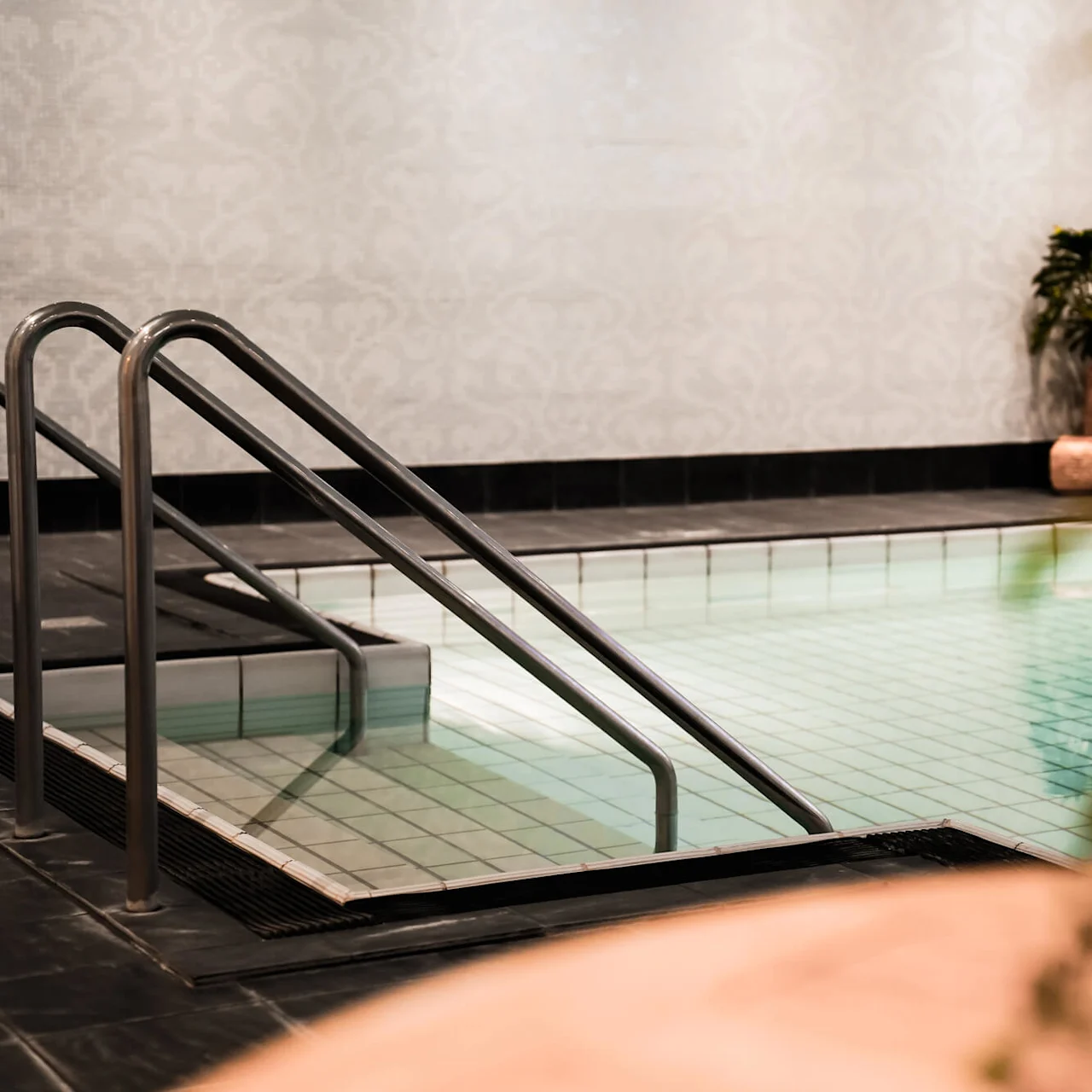 Rostfria stålhandtag ramar in en lugn inomhuspool. Krukväxter dekorerar det kaklade området och skapar en fridfull och inbjudande spa-liknande atmosfär.