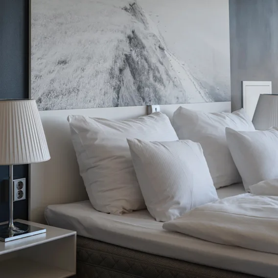 Ett mysigt hotellrum med en bäddad säng, vita lakan och en sänglampa, med ett stort bergskonstverk ovanför sänggaveln.
