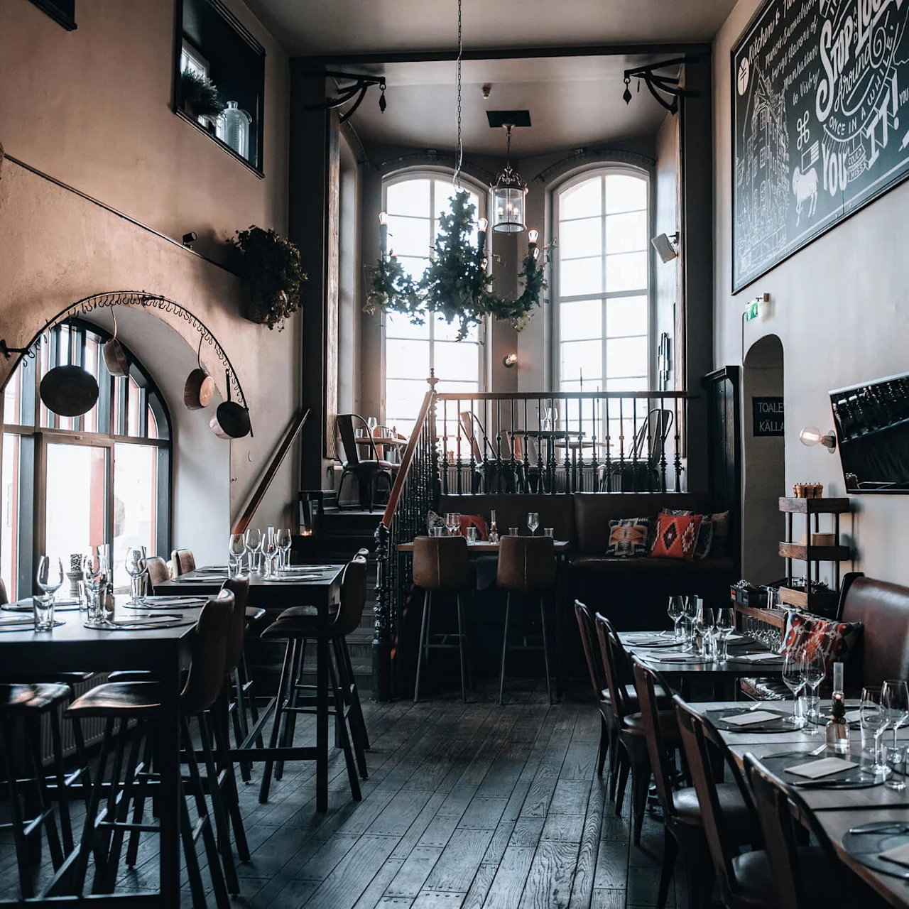 Restaurentinteriør; tomme borde med stoler er pent satt opp med glass og servietter. Øvre seksjon med flere bord, store vinduer lyser opp den koselige, rustikke atmosfæren. Veggkunst og grønne dekorasjoner skaper sjarm.