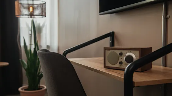 Pieni radio seisoo puupöydällä, jossa on metallituet. Lähellä on viherkasvi ikkunan vieressä, lattiavalaisimen luomassa kodikkaassa huoneessa.