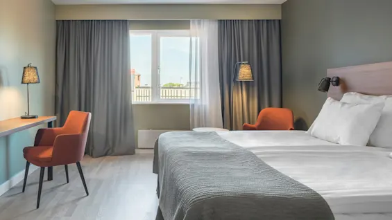 En snyggt bäddad hotellsäng med vita lakan och en grå filt står vid ett fönster, flankerad av två orange stolar; ett skrivbord med lampa finns i närheten.