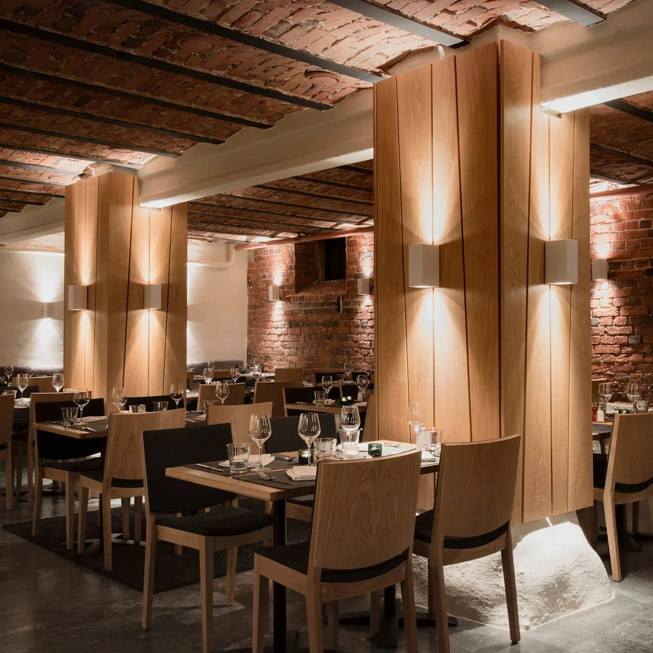 Borde med glas og servietter, omgivet af træstole, fylder en varmt oplyst restaurant med eksponerede murstensvægge og træpaneler, hvilket skaber en intim middagstemning.