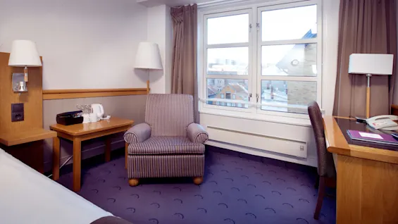 En stribet lænestol står ved et vindue i et ordentligt hotelværelse med sofabord, lampe og seng, med udsigt over bytage.