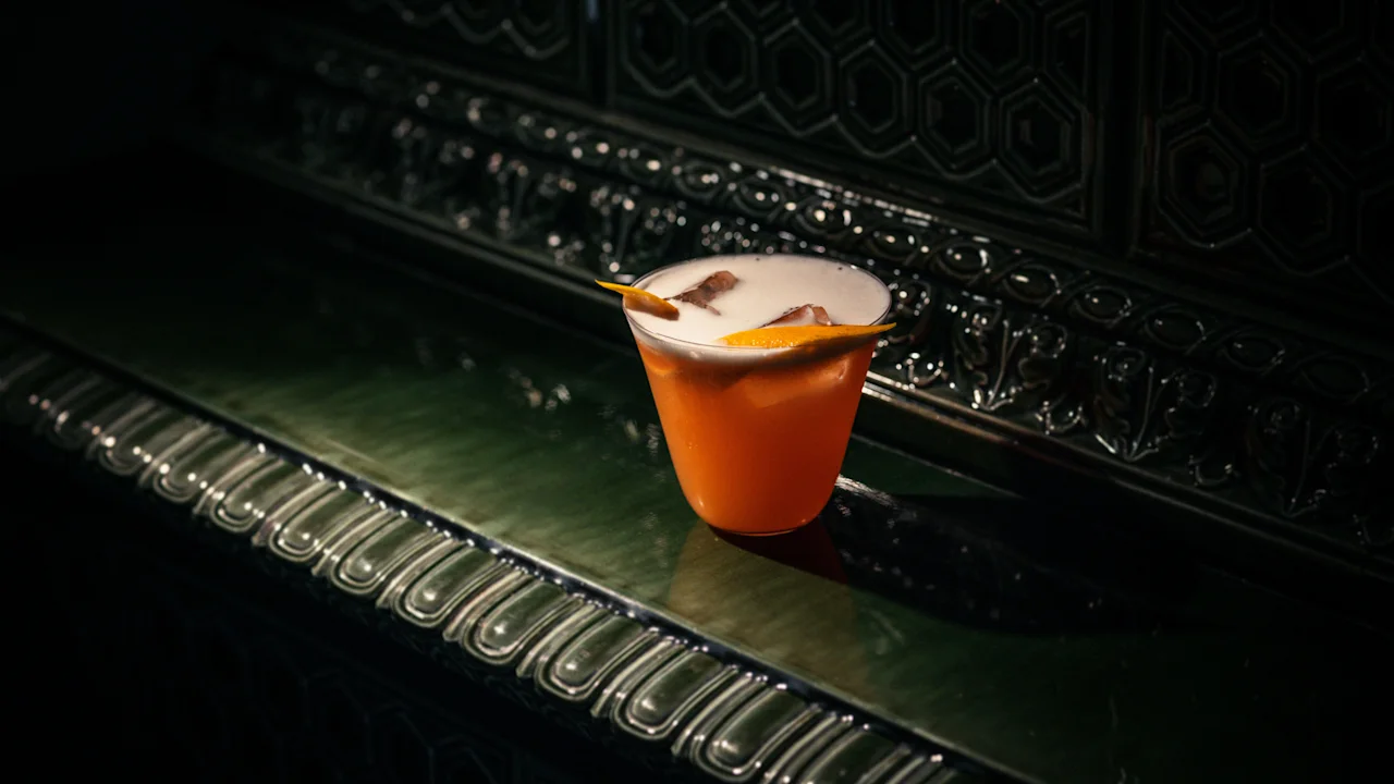 A vibrant cocktail with foam and an orange garnish, set against a dark, ornate background at Hotel Kämp – Kämp Bar.