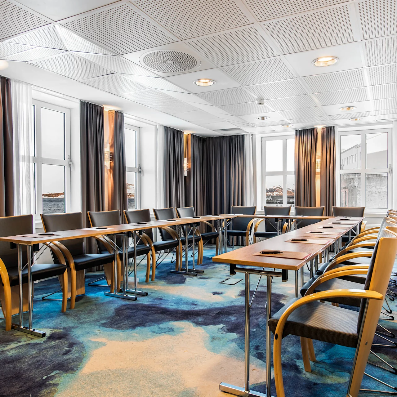 Konferencelokale med arrangerede borde og stole, badet i sollys fra store vinduer. Rummets blå mønstrede tæppe komplementerer dets moderne grå møbler og hvide perforerede loftpaneler.