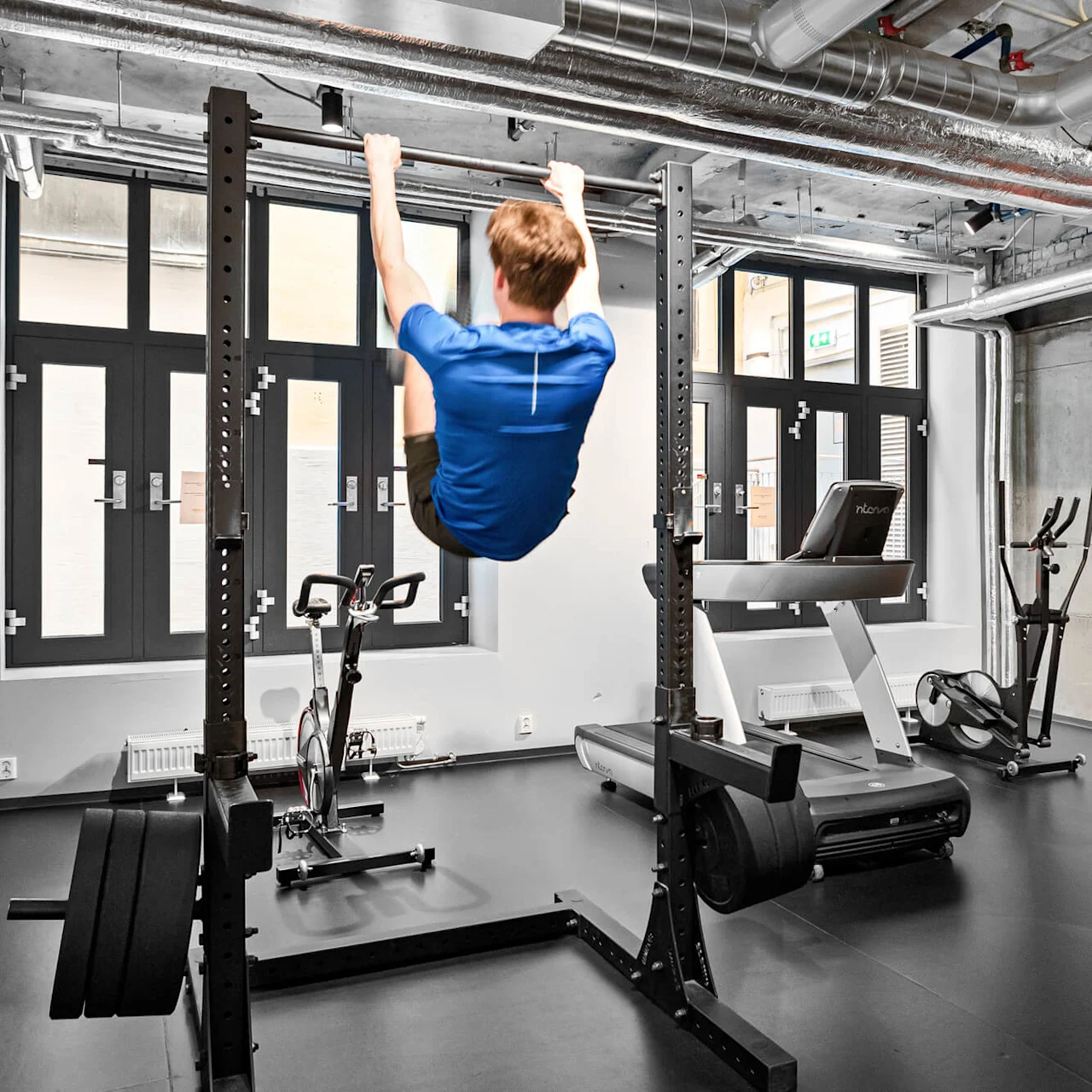 En person i blå tröja gör pull-ups i ett gym. I närheten finns träningscyklar, ett löpband och industriella designelement.