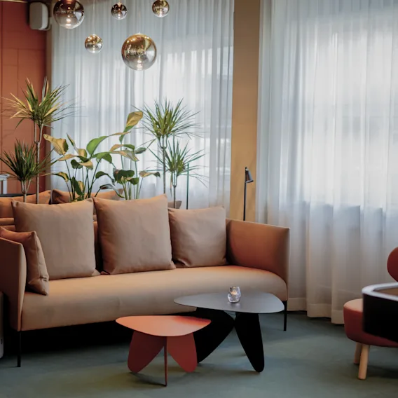 En beige soffa med kuddar står i ett modernt vardagsrum omgiven av växter och dekorativ belysning. Soffbord och en stol bidrar till den mysiga, eleganta atmosfären.