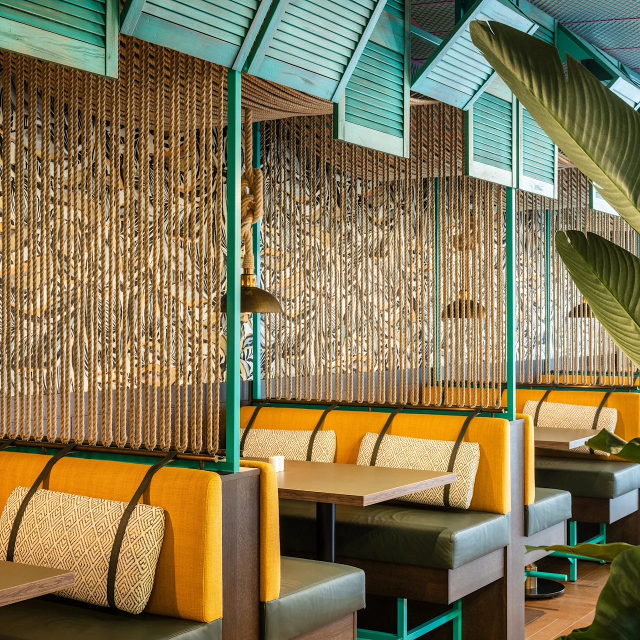 Colorful booths with yellow cushions and patterned pillows arranged in a restaurant. The space features rope partitions, tropical plant decor, and teal ceiling panels, creating a vibrant, cozy atmosphere.