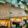 Farverige båse med gule puder og mønstrede puder arrangeret i en restaurant. Rummet har rebopdelere, tropisk planteindretning og teal farvede loftspaneler, der skaber en livlig, hyggelig atmosfære.