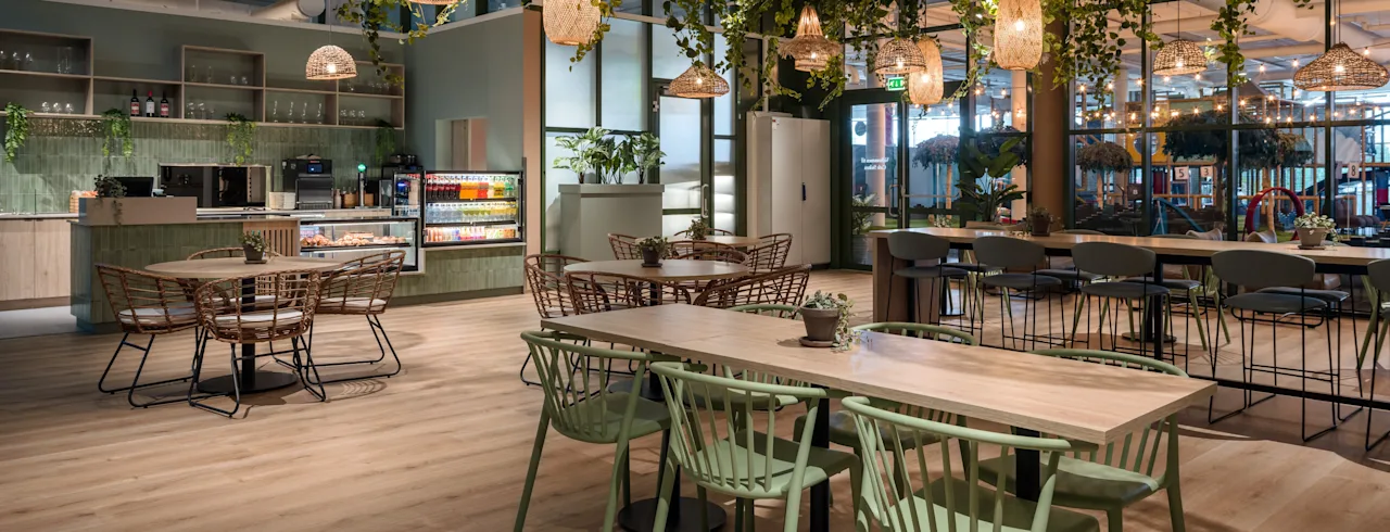 Bord og stoler er arrangert i en stilfull kafé, dekorert med hengende planter og varme pendellamper. Kaféens grønne detaljer og tregulv skaper en hyggelig, innbydende atmosfære.