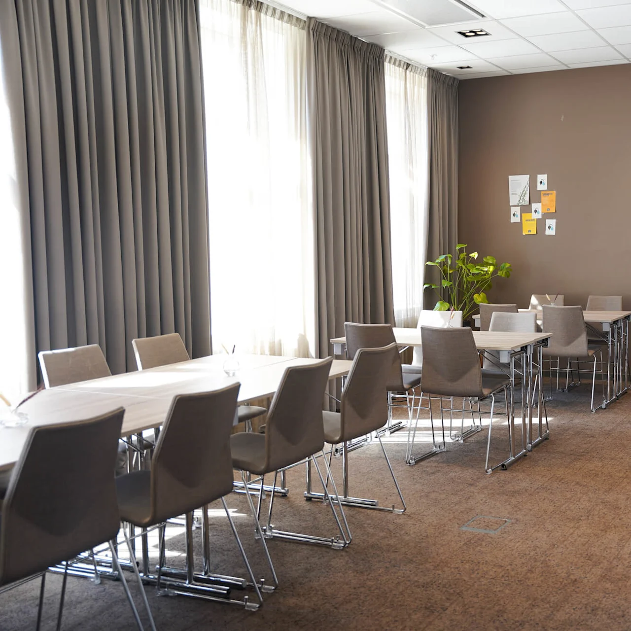 Et konferanserom med ryddig oppsatte bord og stoler, noe glass på bordene, og lukkede gardiner som slipper inn mykt lys. Veggene er dekorert med noen innrammede oppslag.