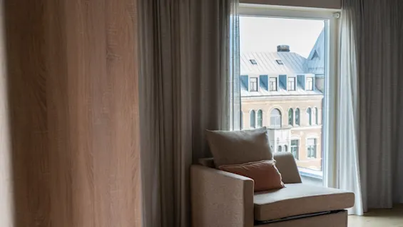 En koselig beige lenestol med puter står ved et stort vindu, med utsikt over en tradisjonell mursteinbygning, omgitt av lysegrå gardiner.