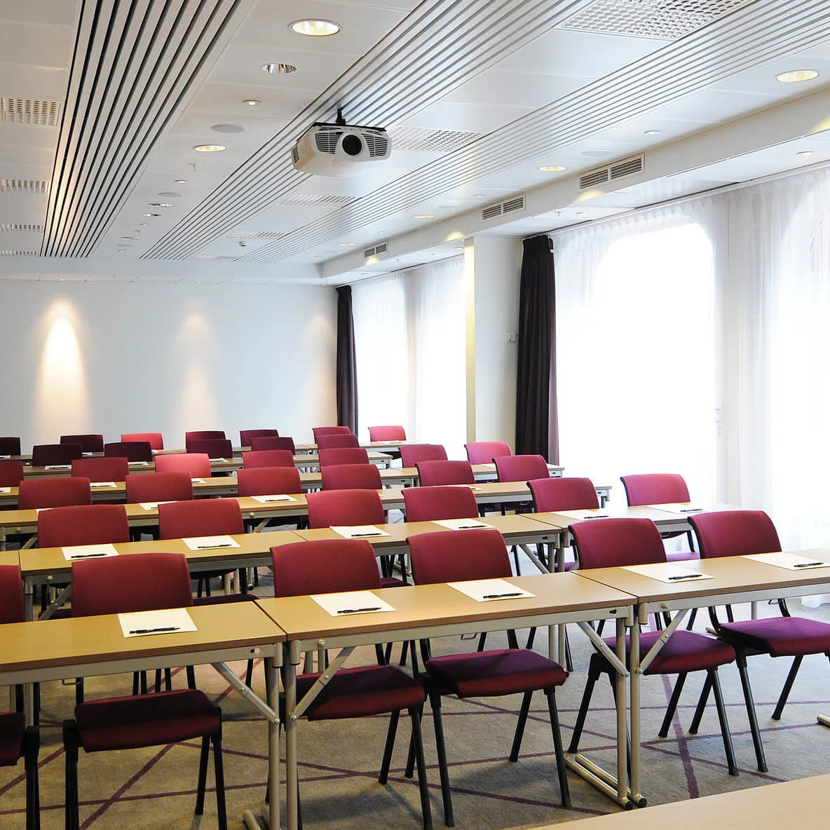 Empty conference room with rows of tables and red chairs, notepads and pens placed on tables, bright lighting, and window curtains partially closed.