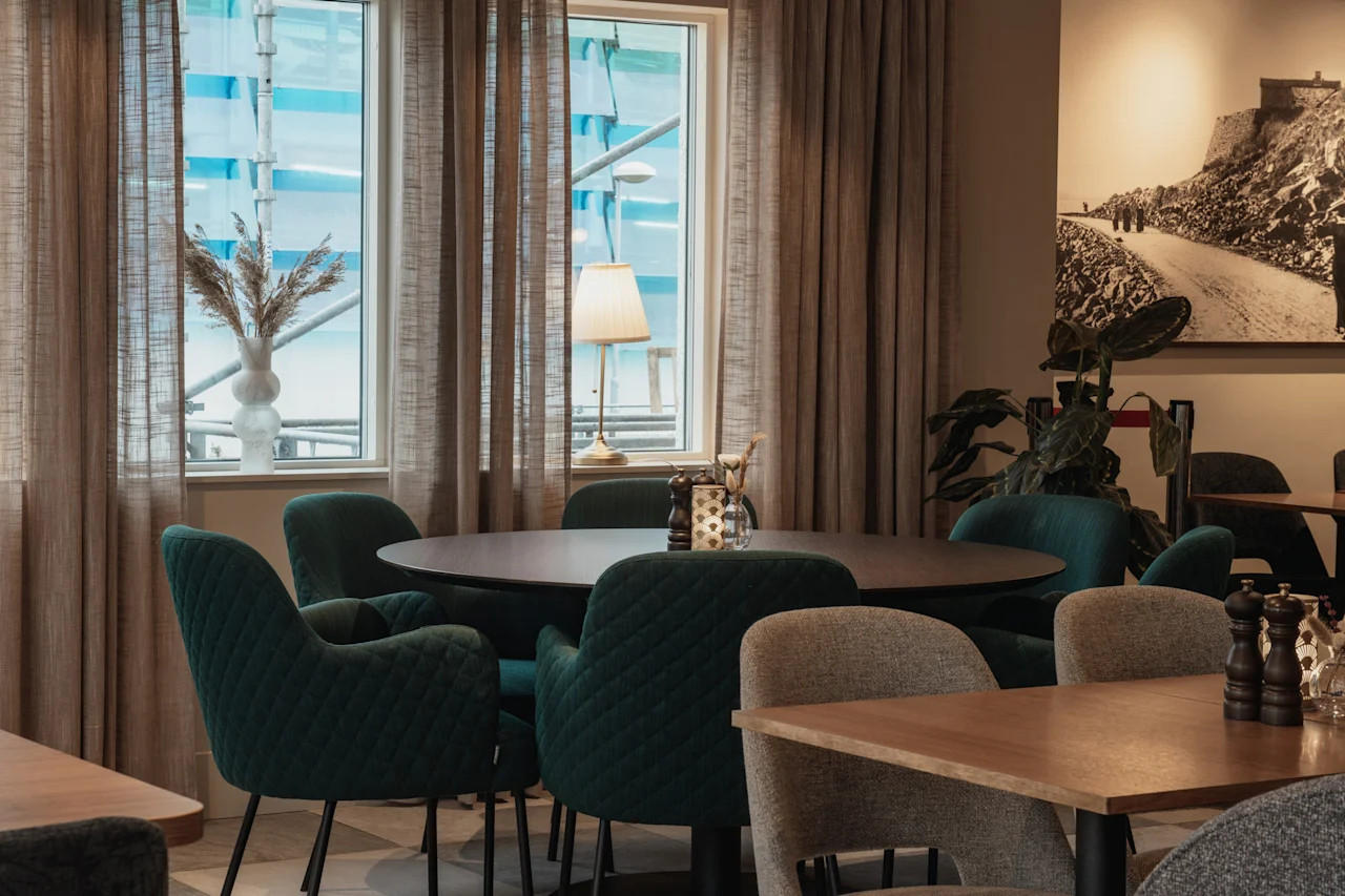 Et komfortabelt spiseområde med runde borde, plysbetrukne stole og store vinduer med flydende gardiner. Et naturskønt kunstværk hænger på væggen.