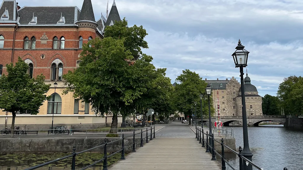 A scenic wooden boardwalk in Örebro, Sweden, alongside a canal with lily pads, leading towards historic buildings under a cloudy sky.